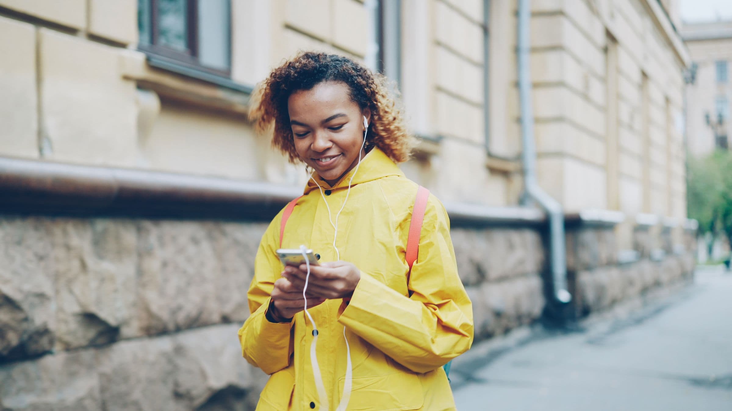 A woman in a yellow raincoat listening to her phone through earphones on a city street